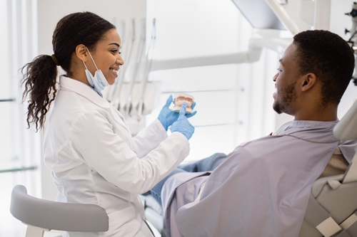 Smiling Black Stomatologist Lady Consulting Male Patient In Clinic, Holding Plastic Jaw