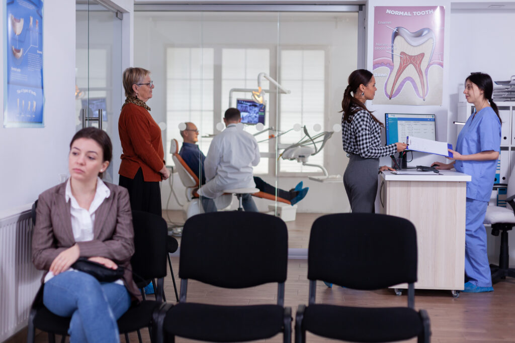 Patients waiting in a dental clinic reception area while a woman checks in at the front desk, illustrating the dental insurance waiting period before treatment.